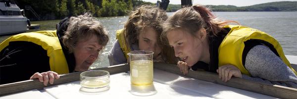 Students and teacher examine a phytoplankton sample from the lake. Photo: Lake Champlain Sea Grant Two students and a teacher in yellow life jackets examine a beaker holding a water sample near a lake